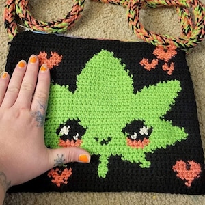 May include: A black crocheted bag with a green marijuana leaf design and orange hearts. The bag has a black and orange braided strap.