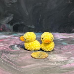 May include: Two small yellow fuzzy duck figurines with black eyes and orange beaks. The ducks are sitting on a purple and gray marbled surface.