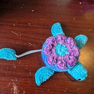 May include: A blue and purple crocheted turtle measuring tape. The turtle has a flower-shaped shell and a long tail that is a measuring tape.