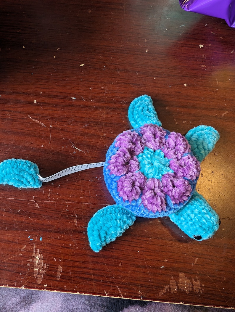 May include: A blue and purple crocheted turtle measuring tape. The turtle has a flower-shaped shell and a long tail that is a measuring tape.