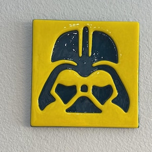 Puede incluir: Un azulejo cuadrado amarillo con una silueta azul oscuro de la cara de Darth Vader.