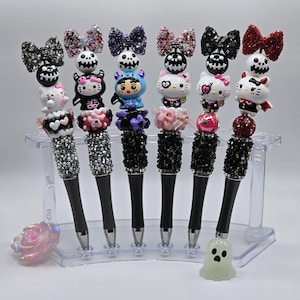 May include: A collection of decorative pens with black ink. Each pen features a unique design, including rhinestone-covered barrels and toppers with skulls, bows, and cartoon characters. The pens are displayed in a clear plastic holder.