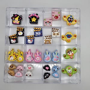 May include: A clear plastic organizer filled with various colorful, cartoon-style charms. The charms include houses, film clapperboards, guitars, clouds, and cactus figures. The color palette includes yellow, pink, blue, and brown, with black and white accents.