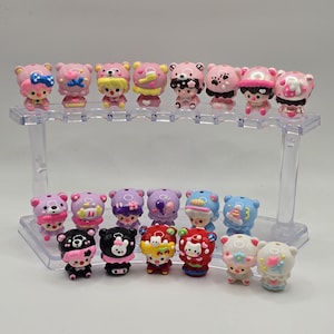 Puede incluir: Una colección de figuras de osos en miniatura en varios colores, incluyendo rosa, morado, negro y blanco, exhibidas en un soporte transparente de dos niveles. Cada oso tiene detalles únicos como lazos, sombreros y elementos decorativos.