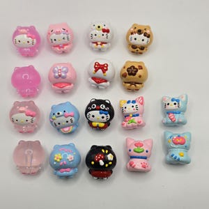 Puede incluir: Una colección de pequeñas figuras de gato de colores en varios diseños. Las figuras presentan diferentes atuendos, lazos y patrones, incluyendo rosa, azul, blanco y marrón. Algunas tienen detalles florales y de hongos.