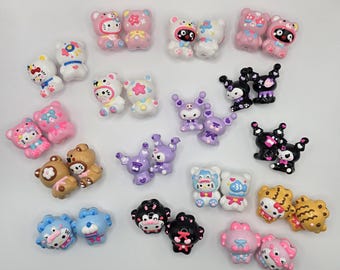 Puede incluir: Una colección de pequeñas figuras con forma de oso de colores, en tonos rosa, azul, blanco, marrón y negro. Algunas figuras tienen caras de personajes y elementos decorativos. Las figuras están dispuestas sobre una superficie blanca.