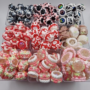 Puede incluir: Una colección de cuentas coloridas con varios diseños, incluyendo florales, lunares y abstractos. Las cuentas están dispuestas en recipientes de plástico transparente, mostrando una variedad de colores como rojo, rosa, blanco y negro.