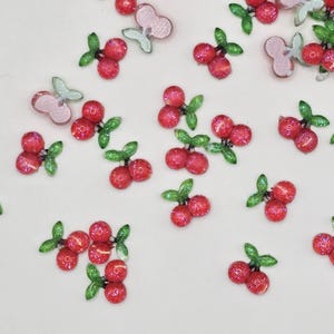 Puede incluir: Una dispersión de adornos de cereza de brillo rojo y verde. Las cerezas son de plástico y tienen un acabado brillante.