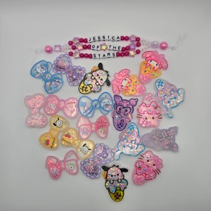 Puede incluir: Una colección de abalorios de resina coloridos, brillantes y brillantes con lazos, corazones y personajes de dibujos animados. Los abalorios están dispuestos sobre un fondo blanco. Los abalorios son de diferentes formas y tamaños. Los abalorios están hechos de resina y tienen un acabado brillante. Los abalorios son muy lindos y serían perfectos para agregar un toque de brillo a cualquier proyecto.