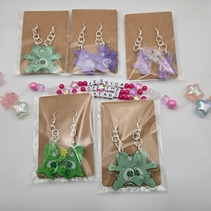 May include: Four pairs of Care Bear earrings on brown card stock. Each pair features two Care Bears holding hands. The Care Bears are green, purple, and blue. The earrings are silver.