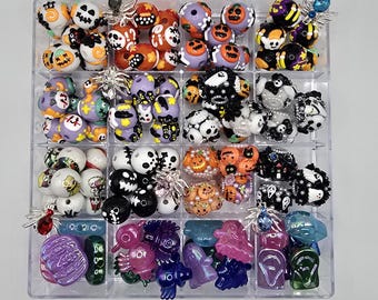 Può includere: Un organizer in plastica trasparente pieno di perline a tema Halloween. Le perline presentano vari disegni, tra cui zucche, teschi, fantasmi e ragni. I colori includono arancione, nero, viola, bianco e verde. Alcune perline hanno ciondoli a forma di ragno argentati.