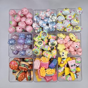 May include: Nine clear plastic containers filled with colorful beads and charms. The beads include pink floral, blue floral, yellow and black striped, purple, and green and yellow designs. The charms include burgers, fries, and ice cream.