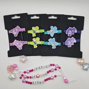May include: A collection of hair clips featuring Care Bear characters in various colors, including purple, green, and blue. Each clip is mounted on a black card. Below, a beaded pacifier clip spells out "JESSICA OF THE STARS" with pink beads and star accents.