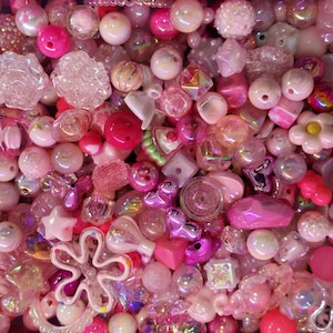 Op de afbeelding: Een close-up van een verzameling roze en witte kralen in verschillende vormen en maten. De kralen omvatten ronde, stervormige, bloemvormige en geometrische vormen. Sommige kralen zijn iriserend, andere ondoorzichtig. Het kleurenpalet wordt gedomineerd door roze en witte tinten.