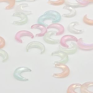 May include: Assortment of iridescent crescent moon shaped charms in various pastel colors, including pink, blue, and green. The charms have a glittery finish.