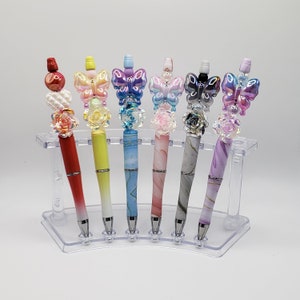 May include: Seven colorful pens with butterfly and flower accents displayed on a clear plastic stand. Each pen features a different color and design, including red, yellow, blue, pink, and purple.