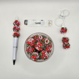 Puede incluir: Un bolígrafo blanco, un llavero y cuentas decorativas con detalles rojos, rosas y verdes. Un tarro contiene varias cuentas decorativas. El texto "16MM" es visible en una etiqueta blanca.