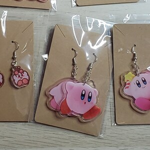 May include: Three pairs of pink and white character earrings. The earrings are on brown card stock with clear plastic packaging. The characters are from the video game Kirby.