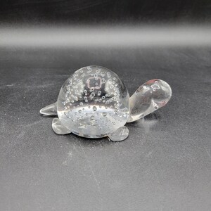 Puede incluir: Una figurita de tortuga de vidrio transparente con una concha llena de burbujas. La tortuga está sentada sobre una superficie negra.