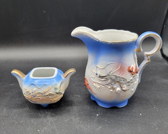 Op de afbeelding: Een blauwe en witte porseleinen roomkan en suikerpot set met een drakenontwerp. De roomkan heeft een gouden handvat en de suikerpot heeft een gouden deksel.