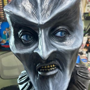 May include: A detailed, close-up of a grotesque mask with a pale, textured surface. The mask features piercing blue eyes, a prominent nose, and a grimacing mouth with yellowed teeth. A ruffled collar is visible.