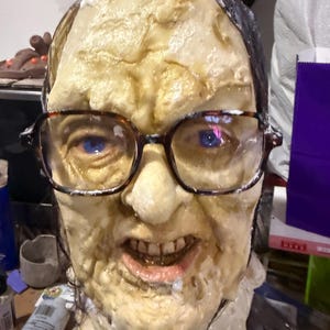 May include: A grotesque, realistic mask of a person with aged skin, wearing glasses. The mask has a pale, yellowish complexion with visible wrinkles and a distorted expression. The glasses have a tortoiseshell pattern.