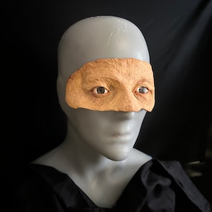 以下が含まれることがあります： マネキンの頭に、老化した皮膚のプロテーゼマスクが目と眉を覆っています。マスクは薄茶色で、詳細なシワとリアルな眼窩があります。マネキンはグレーで、背景は黒です。
