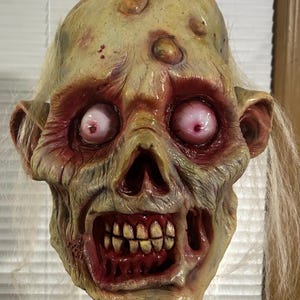 May include: A grotesque zombie mask with a decaying, yellowish-green complexion. The mask features exposed teeth, blood-red eye sockets, and white, stringy hair. Bumps and sores are visible on the forehead, adding to the mask's horrifying appearance. A Halloween costume.