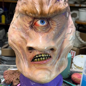 May include: A grotesque mask with a single blue eye, detailed skin texture, and a grimacing mouth revealing yellowed teeth. The mask features a purple neck piece and brown hair, creating a monstrous appearance. The overall color palette is a mix of flesh tones and muted colors.