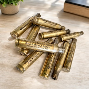 May include: A collection of brass bullet casings engraved with the text UNITED STATES ARMY, SSG AARON J. BARKE, and the dates February 27, 1976 - May 30, 2026. The metallic casings are arranged in a pile, showing various angles of the inscriptions.