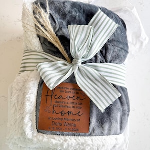 May include: A folded dark gray plush blanket with a white sherpa lining, wrapped in a green and white striped ribbon. A brown leather patch features the text: because someone we love is in Heaven there's a little bit of Heaven in our home.