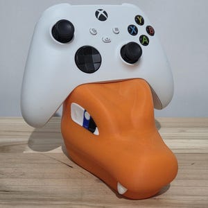 May include: A white Xbox controller sits atop an orange 3D-printed dragon head stand. The controller has black joysticks, coloured lettered buttons, and a directional pad. The dragon stand features a detailed eye and white teeth.