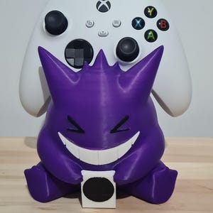 May include: A purple 3D printed Gengar figurine holding a white Xbox controller. The controller is white with black buttons and a black Xbox logo.