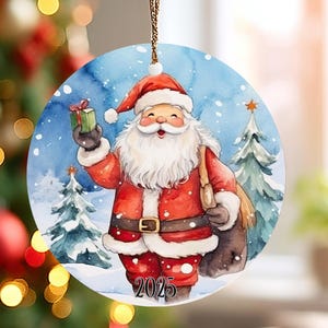 May include: Round Christmas ornament featuring a watercolor illustration of Santa Claus in a red suit and hat, holding a gift. The background shows a snowy scene with pine trees and the year 2025. A gold string is attached for hanging.