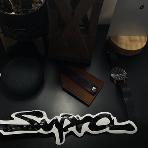May include: A black and white "Supra" sticker with a carbon fiber pattern on a black surface. There is a brown leather key holder with a black strap and a black watch with a silver face on the surface.