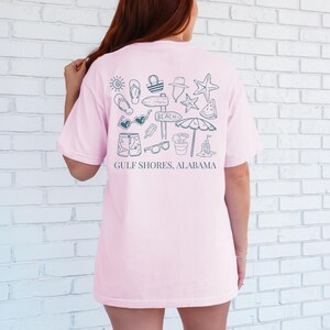 May include: A light pink t-shirt with a beach-themed graphic on the back. The design includes a sun, flip-flops, a beach bag, a hat, a starfish, watermelon, sunglasses, shorts, ice cream, a sign that says "Beach", a sand bucket, a beach umbrella, and a sandcastle. The text "Gulf Shores, Alabama" is below the graphic.