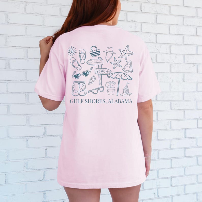 May include: A light pink t-shirt with a beach-themed graphic on the back. The design includes a sun, flip-flops, a beach bag, a hat, a starfish, watermelon, sunglasses, shorts, ice cream, a sign that says "Beach", a sand bucket, a beach umbrella, and a sandcastle. The text "Gulf Shores, Alabama" is below the graphic.