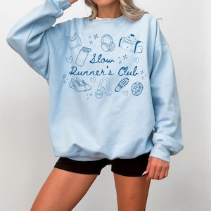 May include: Light blue sweatshirt with a graphic design of running-related items and the text "Slow Runner's Club".