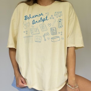May include: A light yellow t-shirt with a blue graphic design that says "Behavior Analyst" and includes various illustrations of toys, a clipboard, a book, and stars.
