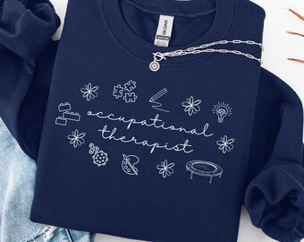 May include: Navy blue sweatshirt with white text that reads "occupational therapist" and white illustrations of objects related to occupational therapy, such as a puzzle, a pencil, a lightbulb, and a trampoline.