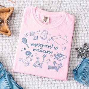 May include: Pink t-shirt with a blue graphic of fitness equipment and the text "movement is medicine".
