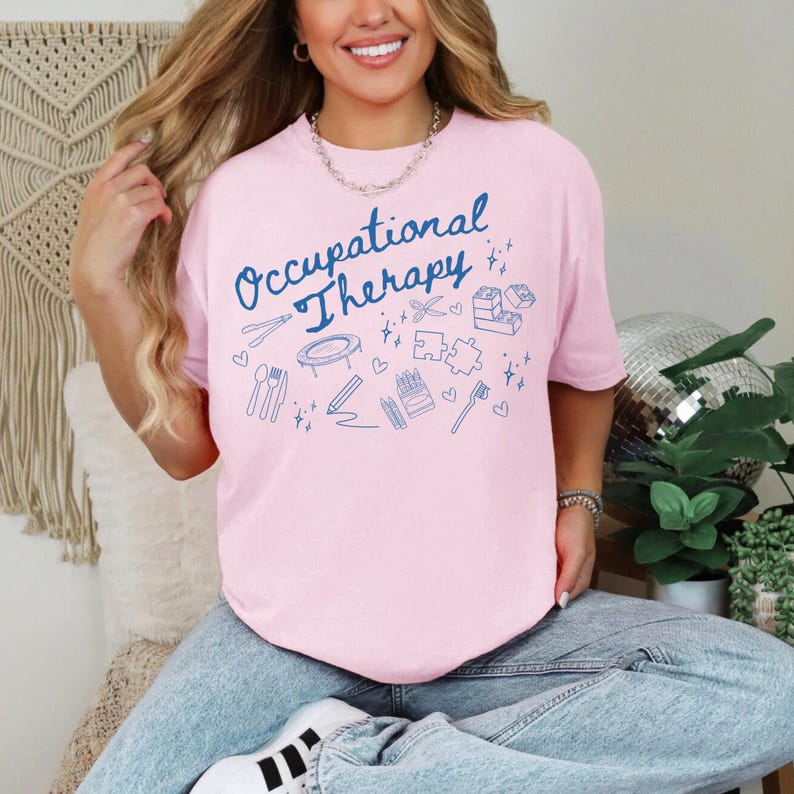 May include: A pink t-shirt with the text "Occupational Therapy" in blue lettering. The shirt is decorated with blue illustrations of various objects related to occupational therapy, including a trampoline, scissors, crayons, a toothbrush, and a puzzle.