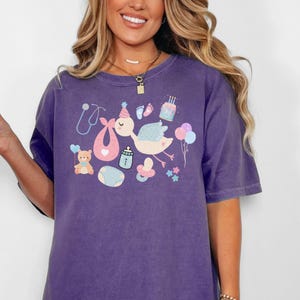 May include: Purple t-shirt with a graphic of a stork carrying a baby, a teddy bear, a pacifier, a bottle, a stethoscope, a bib, and a birthday cake.