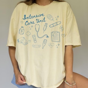 May include: Cream-colored t-shirt with the words "Intensive Care Unit" in blue script, surrounded by blue medical-themed illustrations. The design includes a stethoscope, syringe, and heart rate monitor.