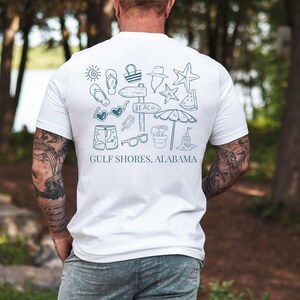 May include: White t-shirt with a teal graphic of beach-themed icons, including sunglasses, a starfish, and a beach sign. The text "GULF SHORES, ALABAMA" is printed below the graphic. The shirt is short-sleeved.