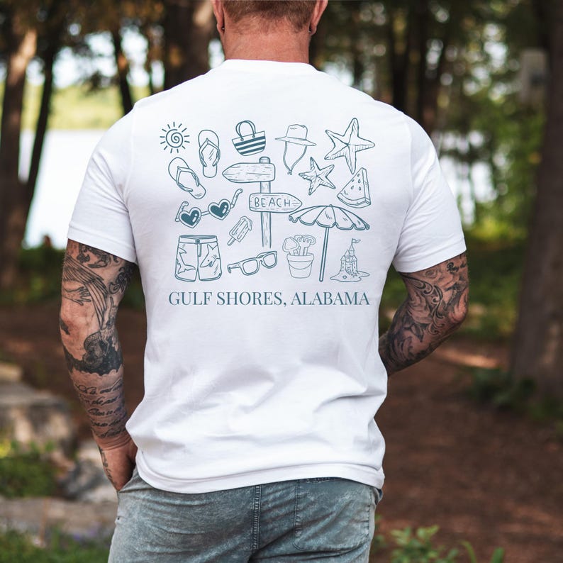 May include: White t-shirt with a teal graphic of beach-themed icons, including sunglasses, a starfish, and a beach sign. The text "GULF SHORES, ALABAMA" is printed below the graphic. The shirt is short-sleeved.