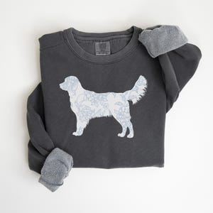 Op de afbeelding: Donkergrijze sweatshirt met een wit honden silhouet gevuld met een blauw bloemenpatroon. De sweatshirt heeft een ronde hals en lange mouwen met grijze fleece manchetten. Het ontwerp is gecentreerd op de voorkant.