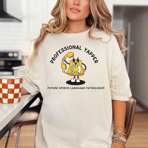 May include: White t-shirt featuring a cartoon illustration of a yellow rotary phone with the text "Professional Yapper" and "Future Speech Language Pathologist".