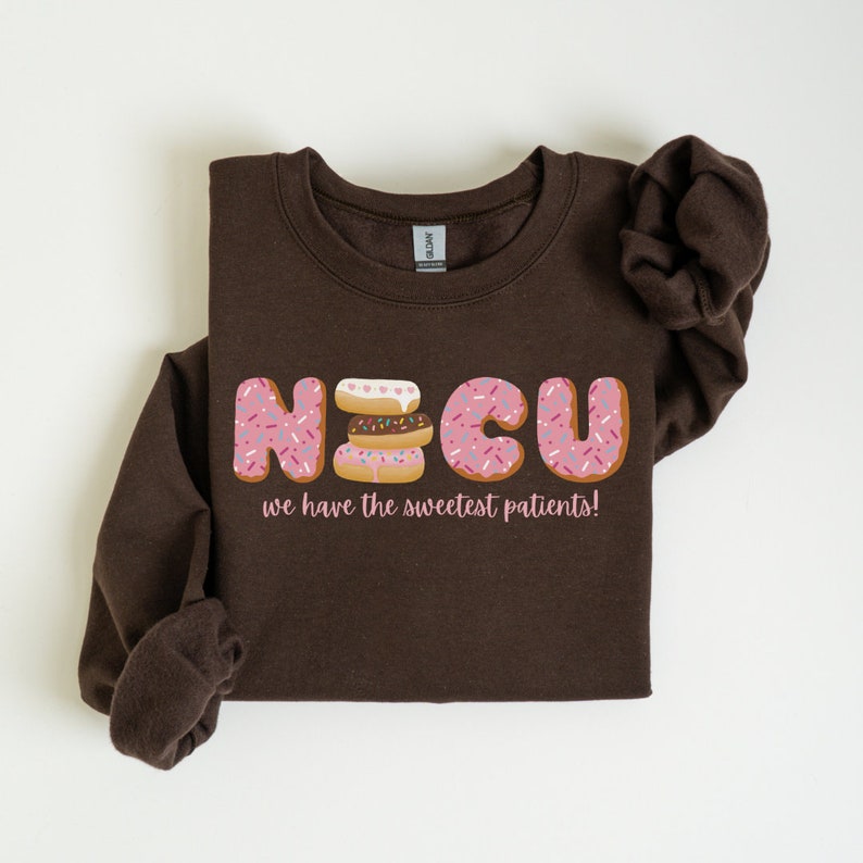 Pode incluir: Camisola de cor castanho com um gr&aacute;fico de donut rosa e branco que soletra "NICU" e o texto "we have the sweetest patients!"