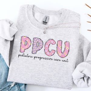 May include: A light gray sweatshirt with the letters "PPCU" in a colorful floral design. The text "pediatric progressive care unit" is printed below the letters. A silver necklace with a small pendant is also visible.